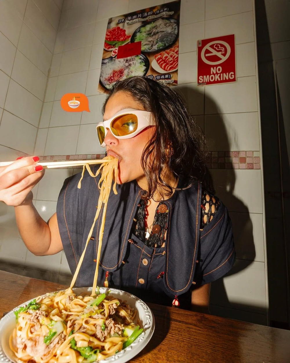 Dunkelhaarige Frau mit sportlicher Sonnenbrille sitzt in asiatischem Restaurant und isst genüsslich Ramen von einem Teller